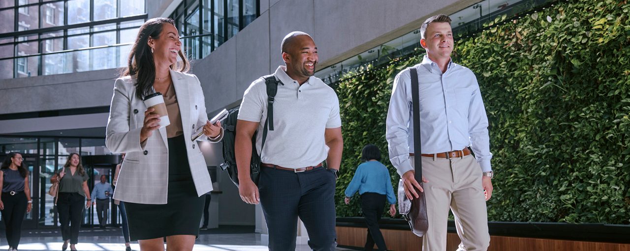 Two male employees and one female walking in a well-lit building lobby with smiles on their faces.