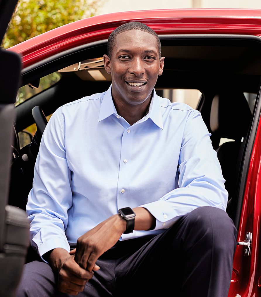 Dark-skinned gentleman with a light blue shirt sitting in a red vehicle and smiling.