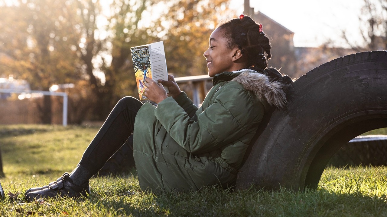 A young lady, in a seated position on green grass, leaning on a big tire and reading a book.