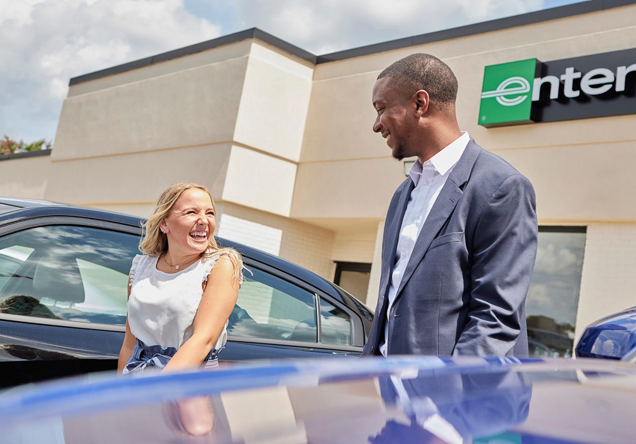 A young man and young woman, both smiling on a sunny day, talk on an Enterprise rental car lot.