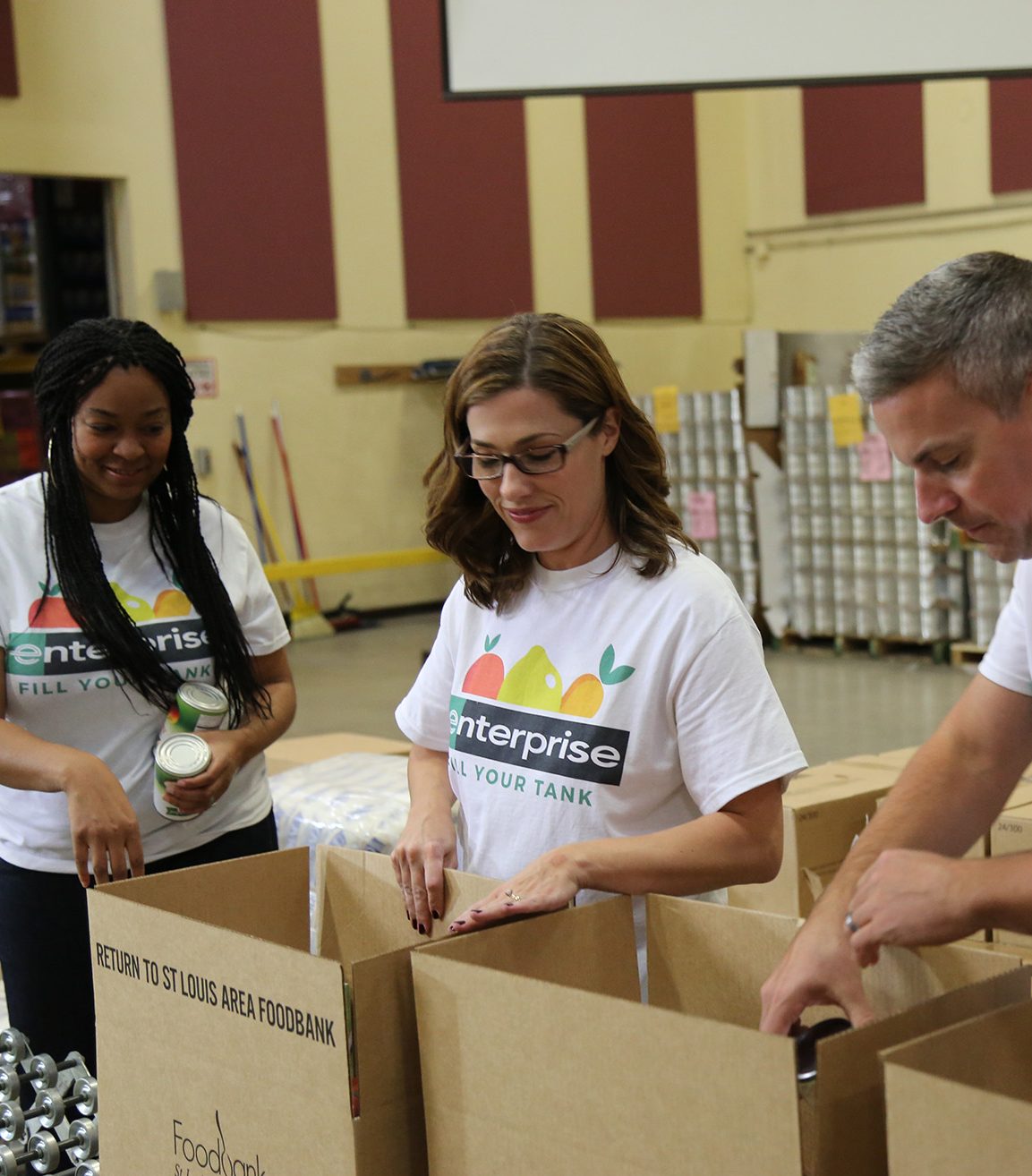 Two female Enterprise employees and one male employee, all wearing white Enterprise Fill Your Tank t-shirts, pack boxes during a community service project.
